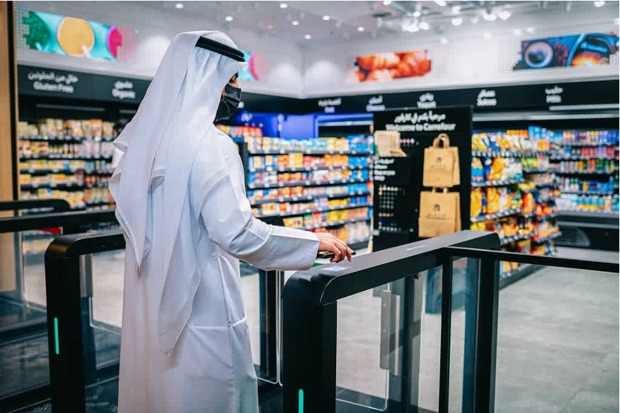 A man in traditional Middle Eastern attire stands at an automated supermarket gate. The store features organized aisles, digital sensors, and a cashierless shopping experience.
