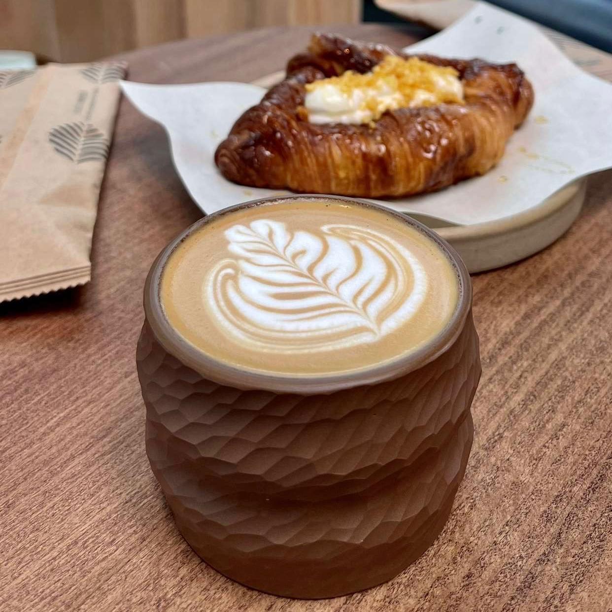 A coffee with a beautiful coffee art on top of it, inside an earthy toned color coffee cup on a wooden table making Bayn an aesthetic café. An earthy looking plate behind it, that has a croissant with cheese, and a packed cutlery.