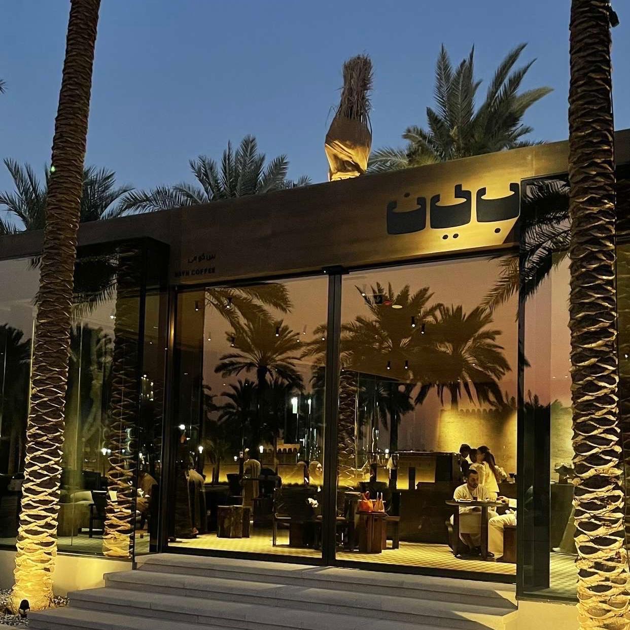 An aesthetic café surrounded by palm trees, an earthy coloured sign with an Arabic word that says Bayn “بين” which means between. Also, glass walls with a reflection of the palm trees and Qasr Al Muwai on it, dim lightning, customers inside the café, and palm trees surrounded with glass walls too.