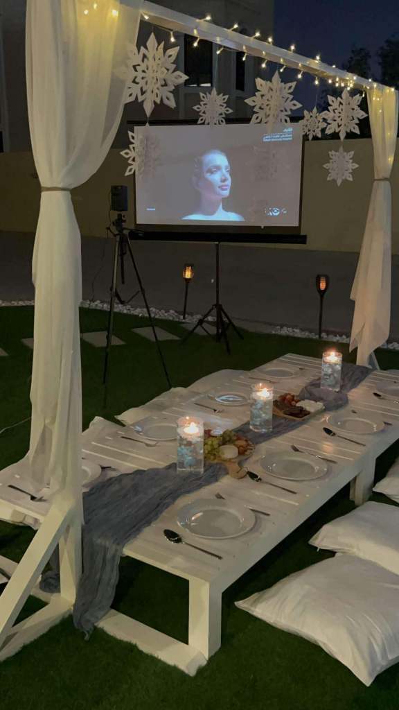 An outdoor home cinema setup with a projector, fairy lights, cozy seating, and a decorated dining table. 