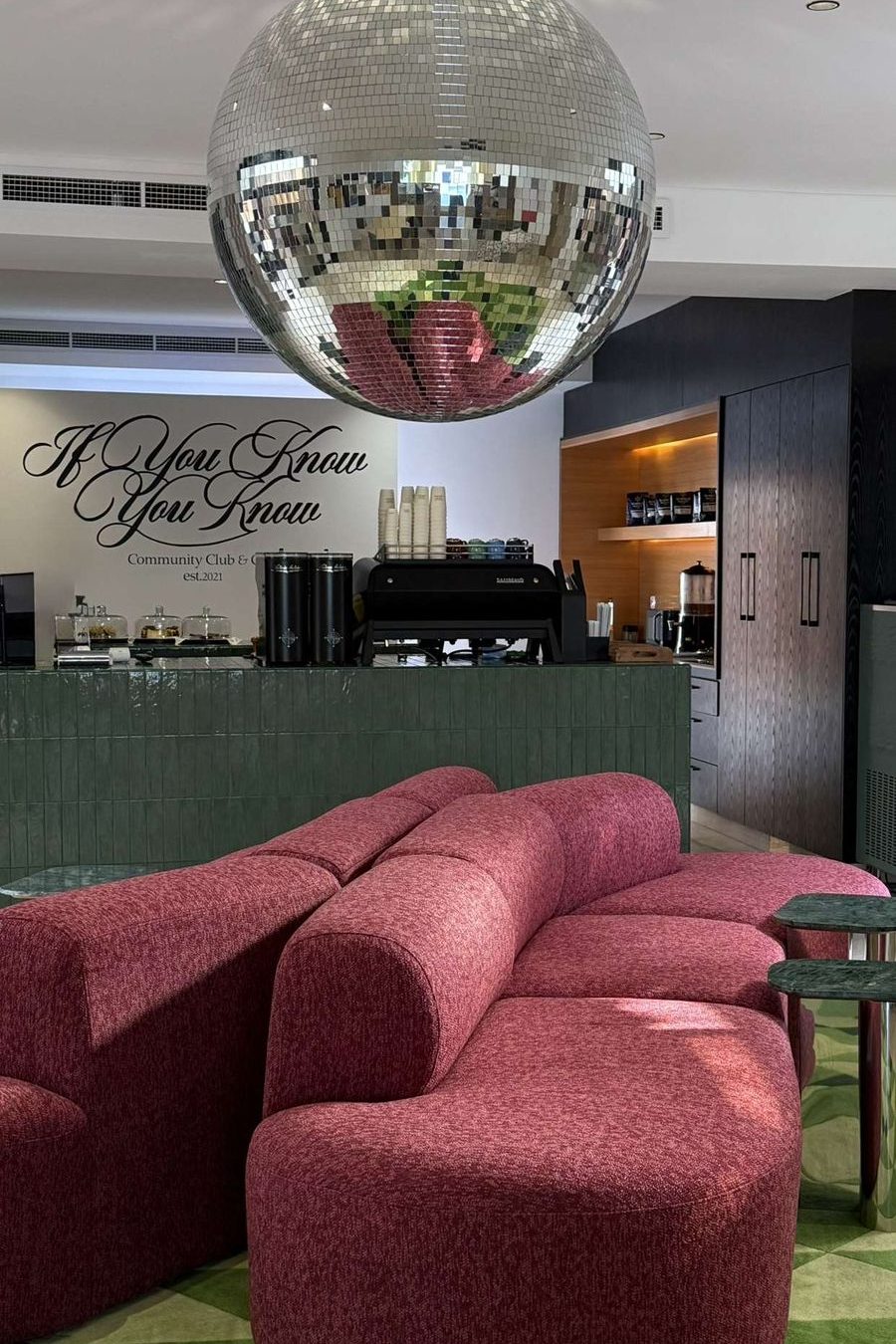 A aesthetic café with a large disco ball, a plush red curved sofa, and a green geometric floor. The counter features a black espresso machine and a sign reading ‘If You Know You Know.