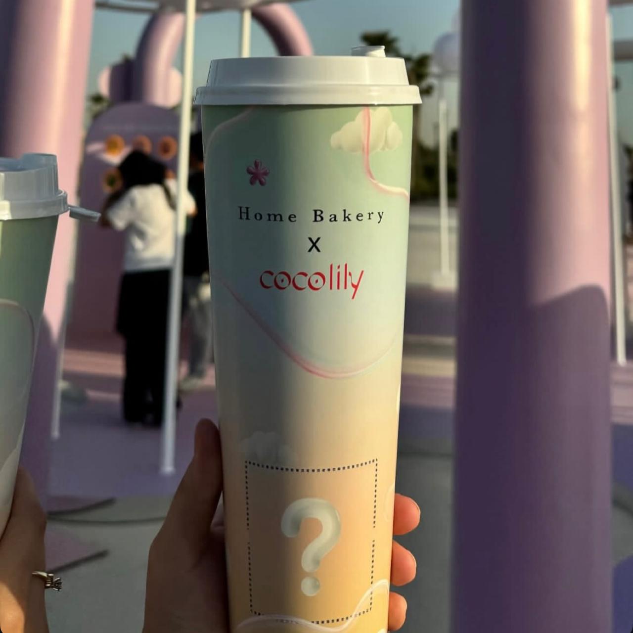 Tall pastel-colored takeaway cup from 'Home Bakery x Cocolily,' part of a mystery collectible series. The cup features a dreamy, aesthetic design with a question mark indicating a surprise item. The background showcases an Instagrammable cafe setting in Dubai, with soft pink and purple decor and people enjoying the space