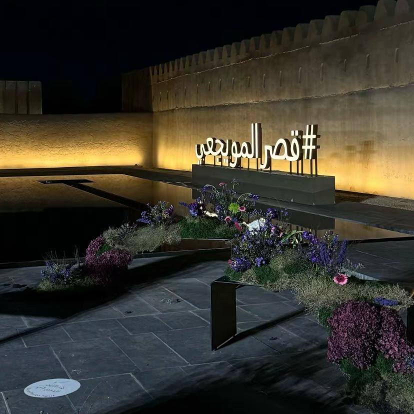 A white sign in Qasr Al Muwaij, that says Qasr Al Muwaiji in Arabic ,floral decoration, and soft dim lightning on the bottom of the wall. A beautiful and strategic location for an instagrammable café, called Bayn Café.